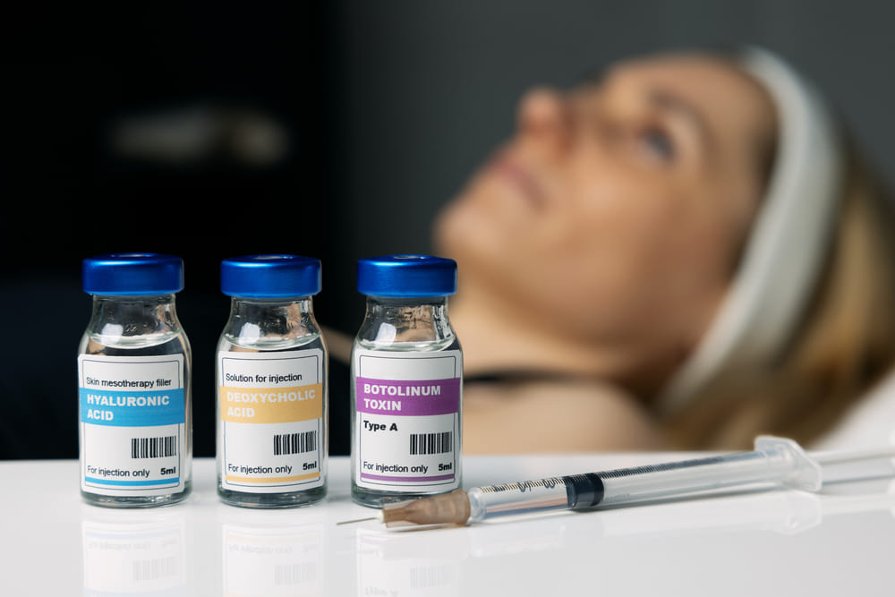 Botox and Dysport vials with syringe in foreground and patient receiving facial treatment in background